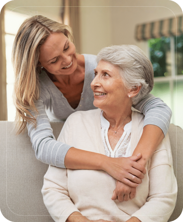 Young woman affectionately hugging senior woman while smiling on a couch.
