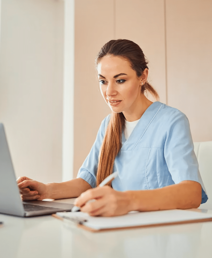 Text Alongside Image Right Woman in blue scrubs working on a laptop at a desk with a clipboard